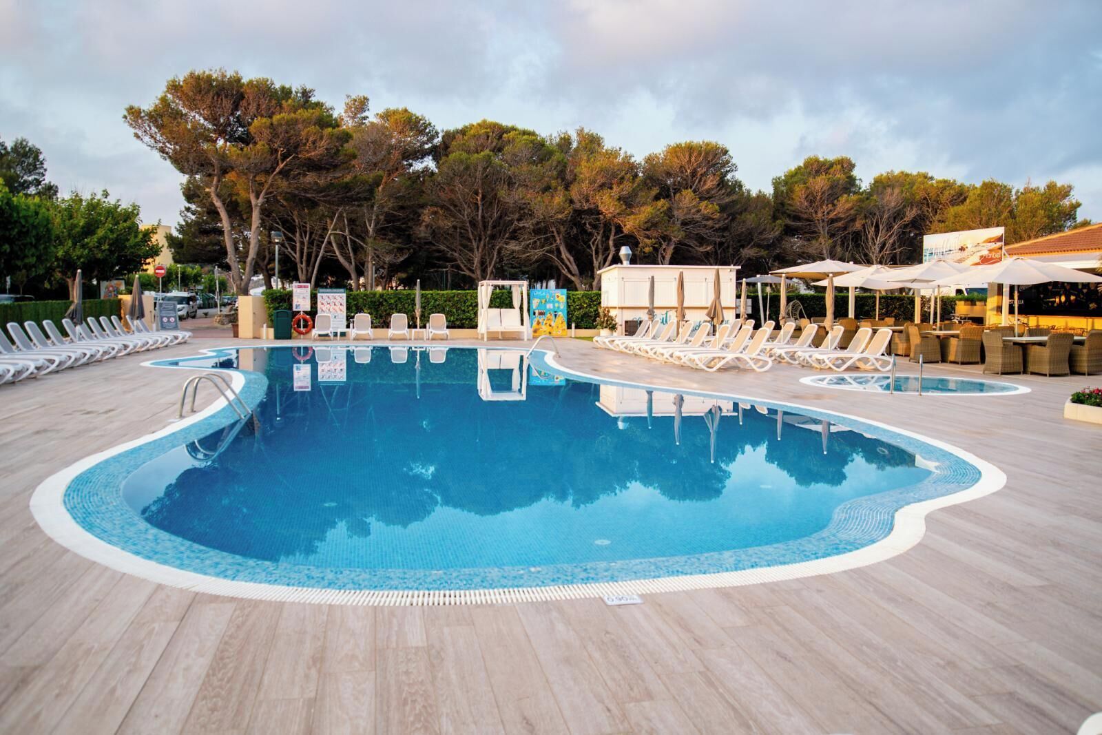 Une piscine entourée d’arbres et d’un bâtiment, avec des chaises longues et des parasols.