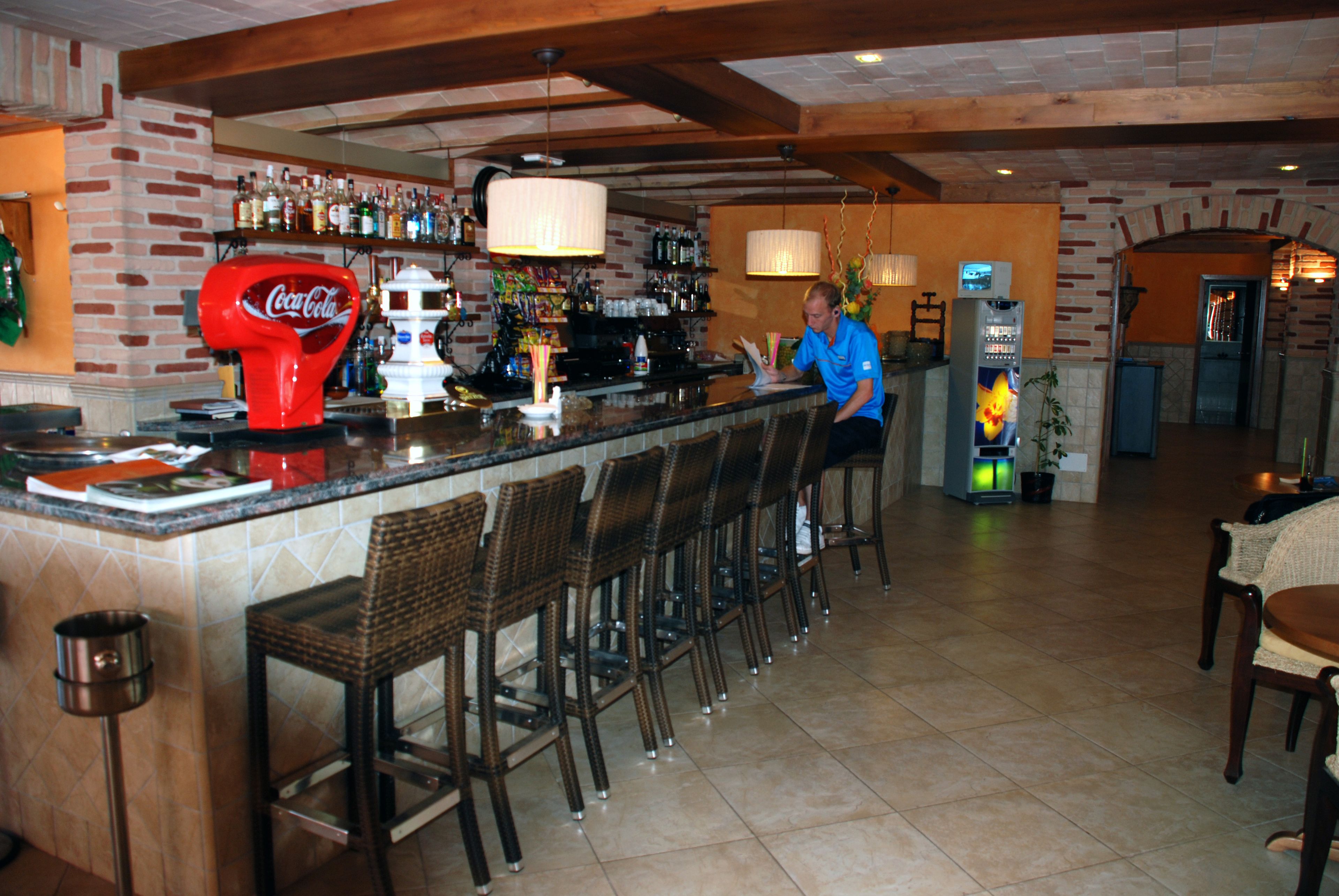 Un espace bar doté d’un distributeur arborant la marque Coca-Cola, d’un barman et d’un coin salon avec des chaises en osier.