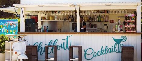 Un bar exterior blanco llamado 'Chillout Cocktail Bar' con una barra que muestra diversas botellas y aperitivos.