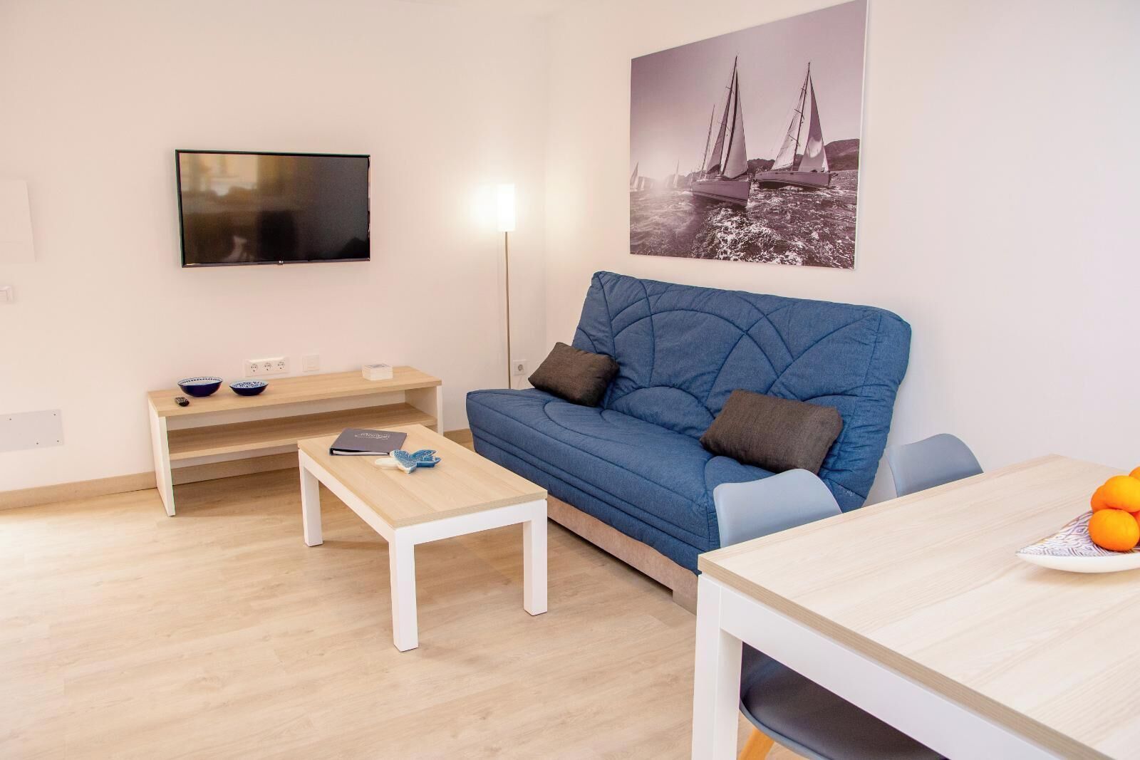 Ein modernes Wohnzimmer mit einem blauen Sofa, einem Holztisch, einem an der Wand befestigten Fernseher und einem Gemälde von Segelbooten.
