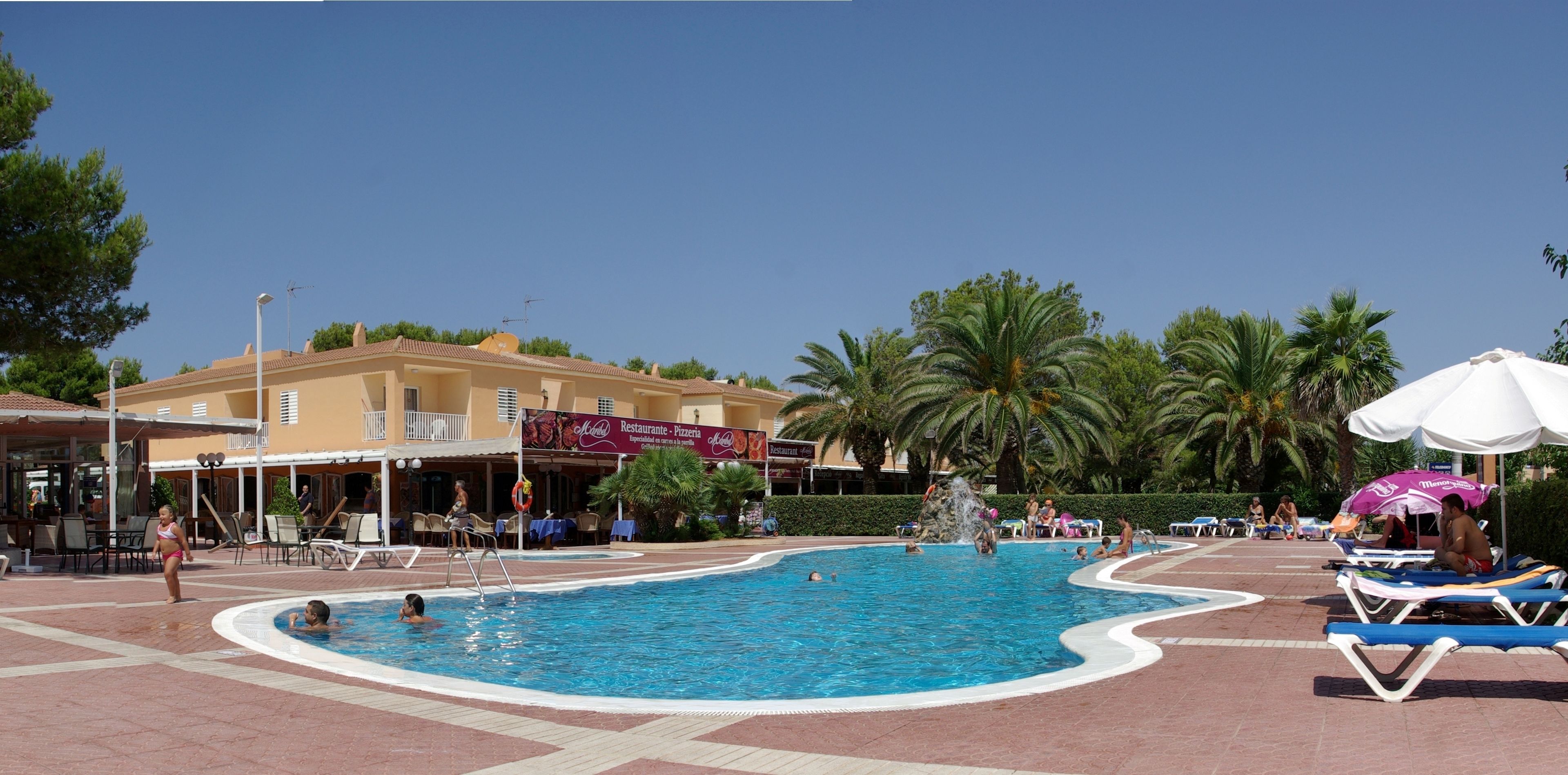 A swimming pool with lounge chairs and umbrellas, a restaurant in the background, and palm trees.