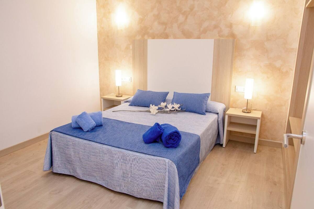 Un lit bien fait, avec une literie et des oreillers bleus, une table de chevet avec une lampe et un luminaire fixé au mur.