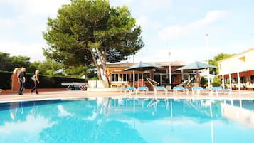 Relax by the pool surrounded by lounge chairs and umbrellas, with a tree providing shade and a nearby pub.