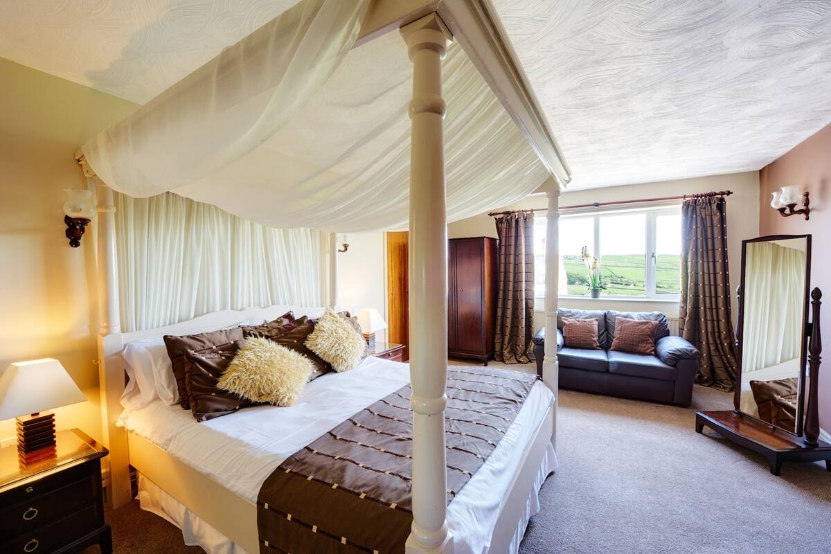 A four-poster bed with a canopy, a bedside table with a lamp, a sofa, and a mirror in a well-lit room with large windows.