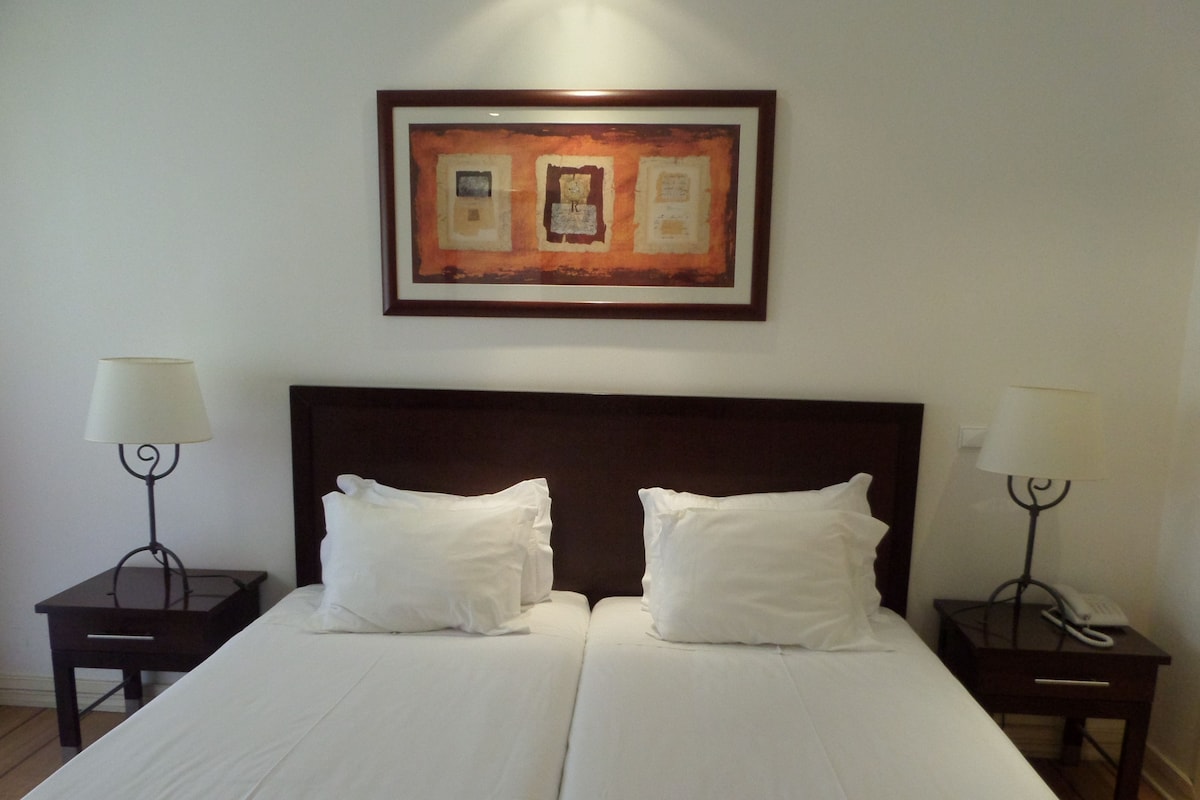 A double bed with white linens, two bedside tables with lamps, and a framed artwork above the bed.