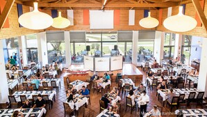 Un restaurante espacioso con techos altos y ventanas grandes, que cuenta con varias mesas y lámparas colgantes.