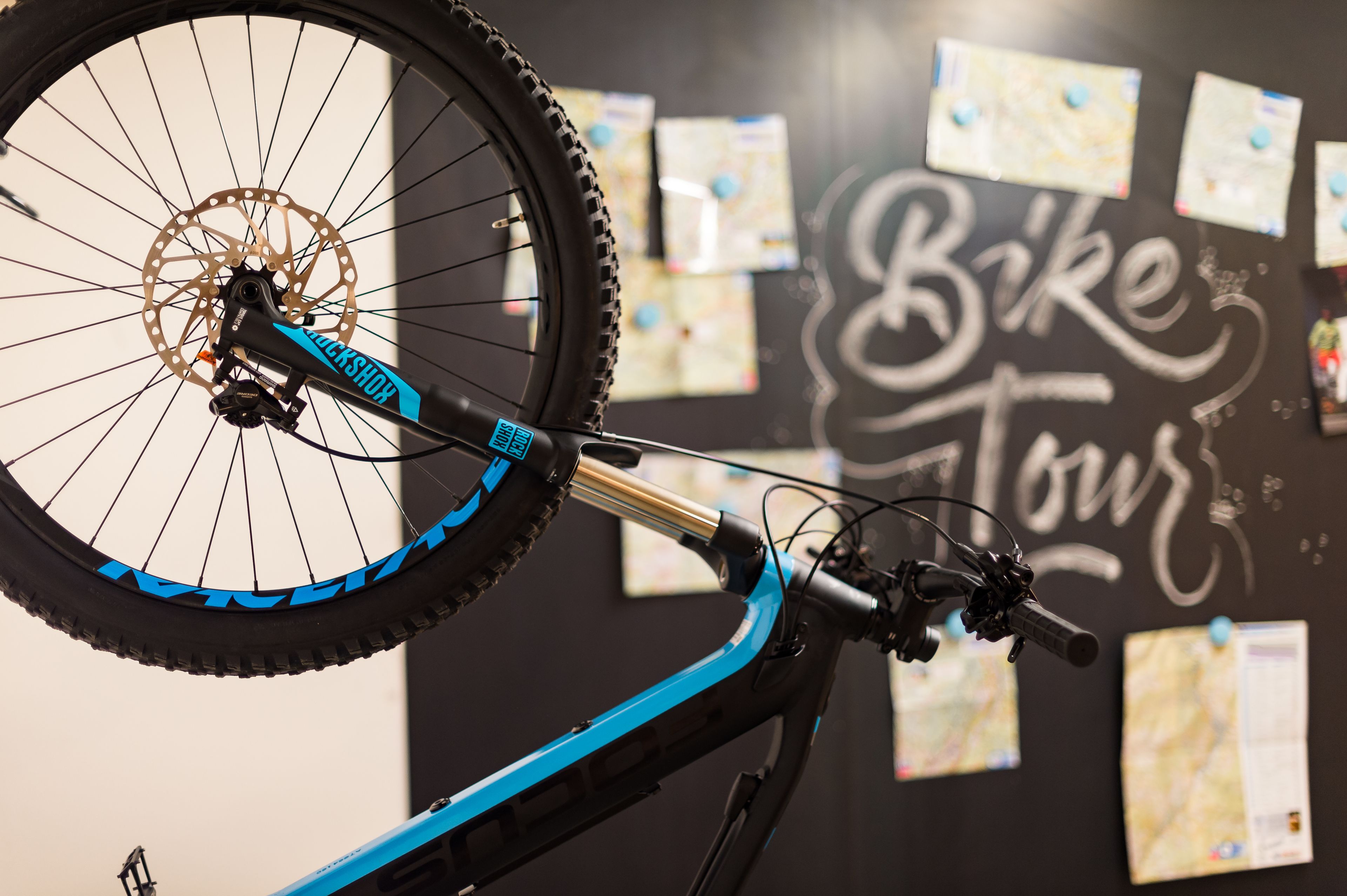 Una bicicletta con freno a disco e telaio blu, posizionata di fronte a un muro con un cartello 'Bike Tour' e mappe affisse.