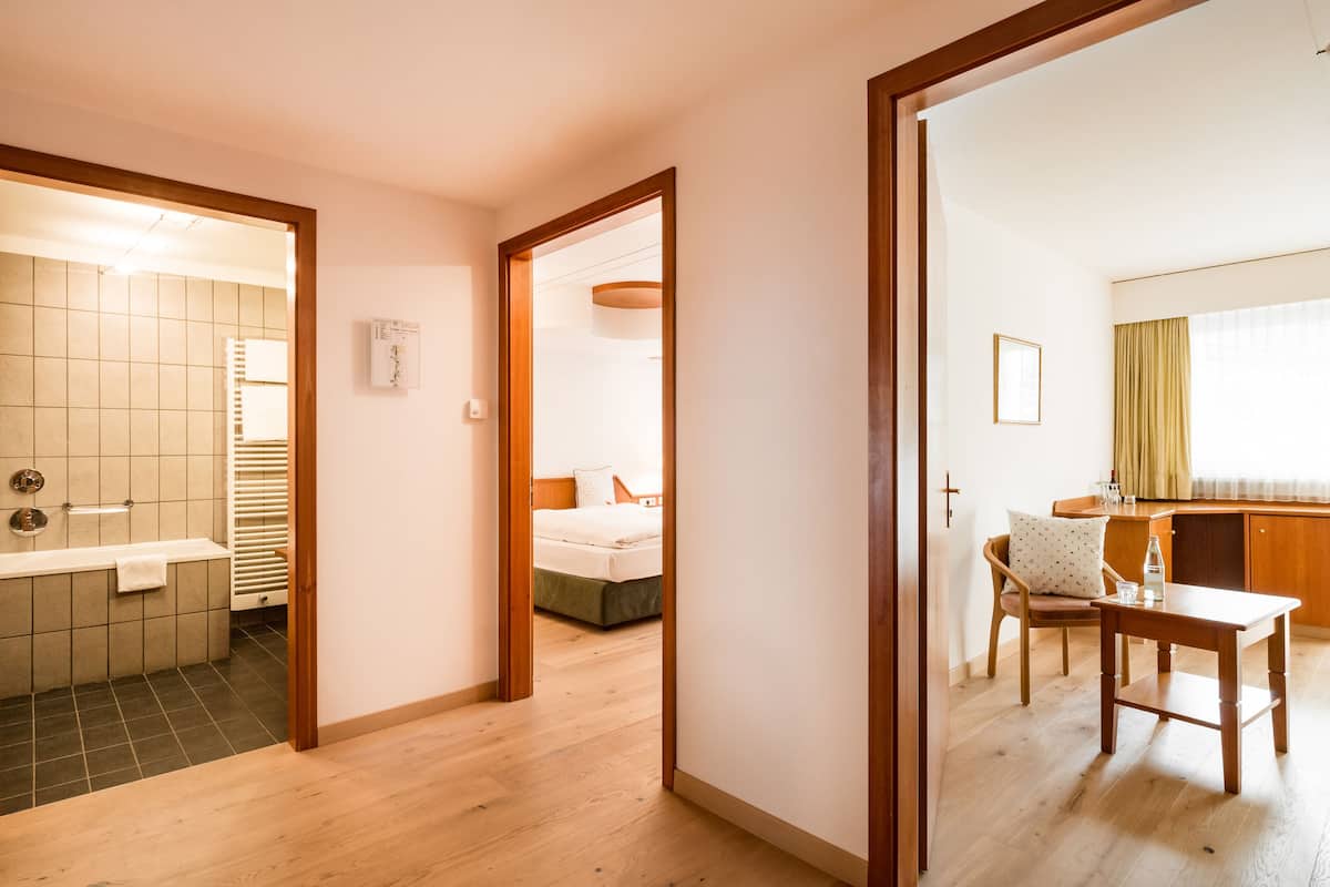 A hotel room with a wooden floor, a bed with a green bedspread, a small table, a chair, and a view of a bathroom with a bathtub and a towel rack.