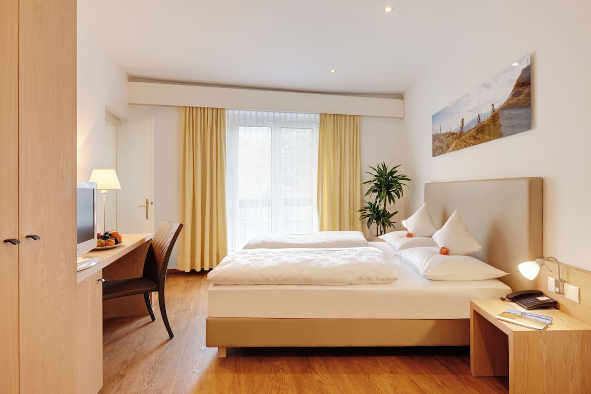 A hotel room with a bed, bedside table, lamp, desk, chair, and a window with curtains.