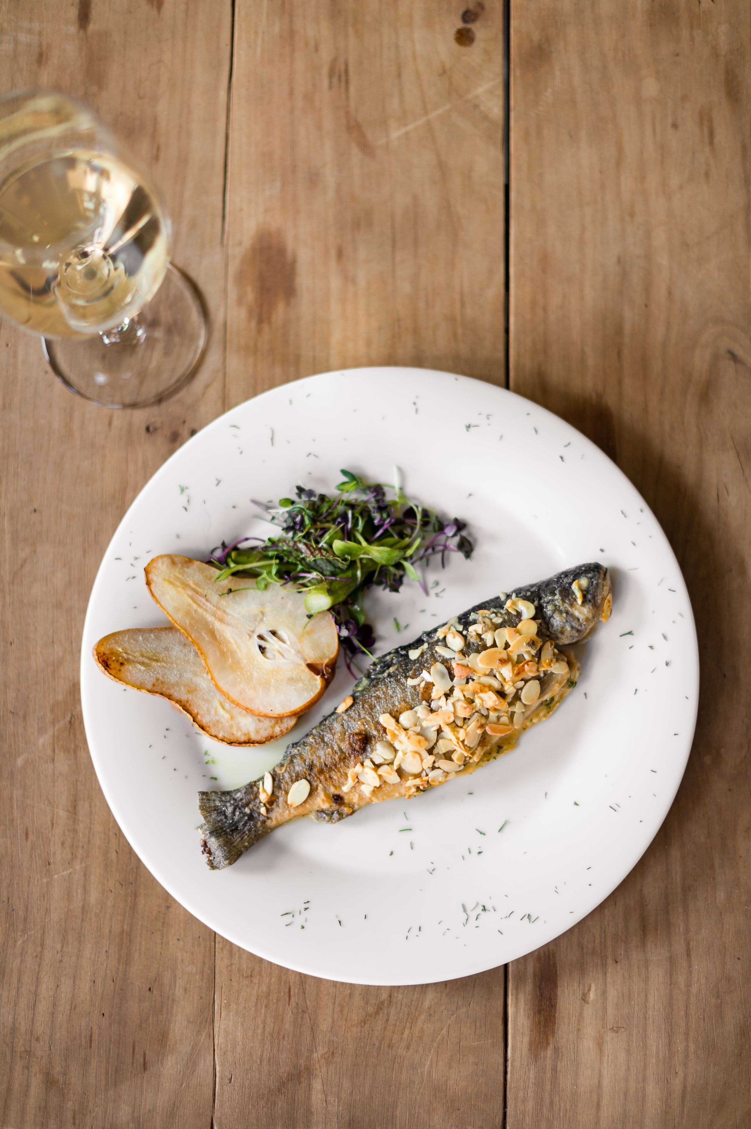 A white plate with a fish, potato slices, and greens on a wooden table.