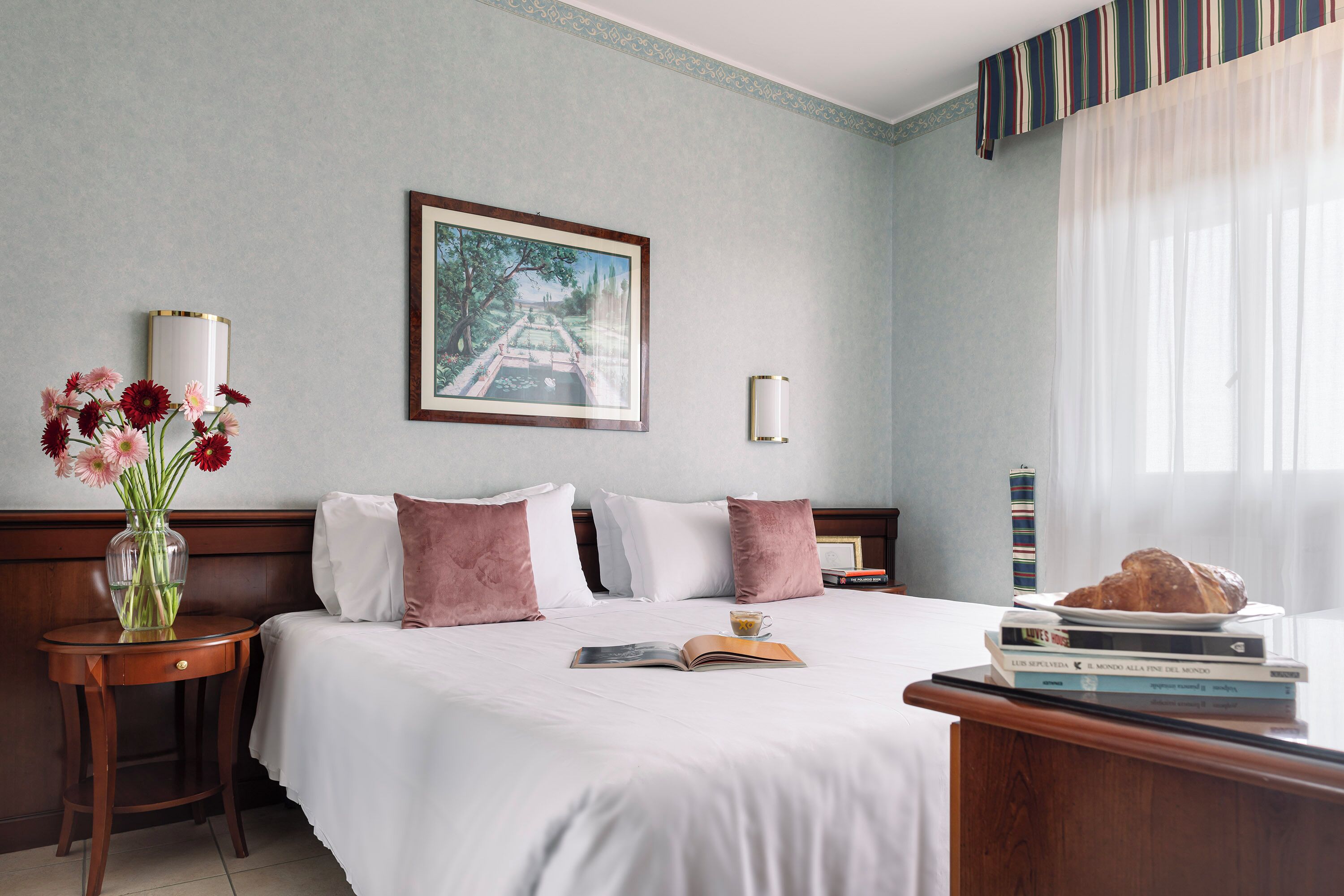 A neatly made bed with white and pink pillows, a vase with flowers, and a stack of books on a bedside table.