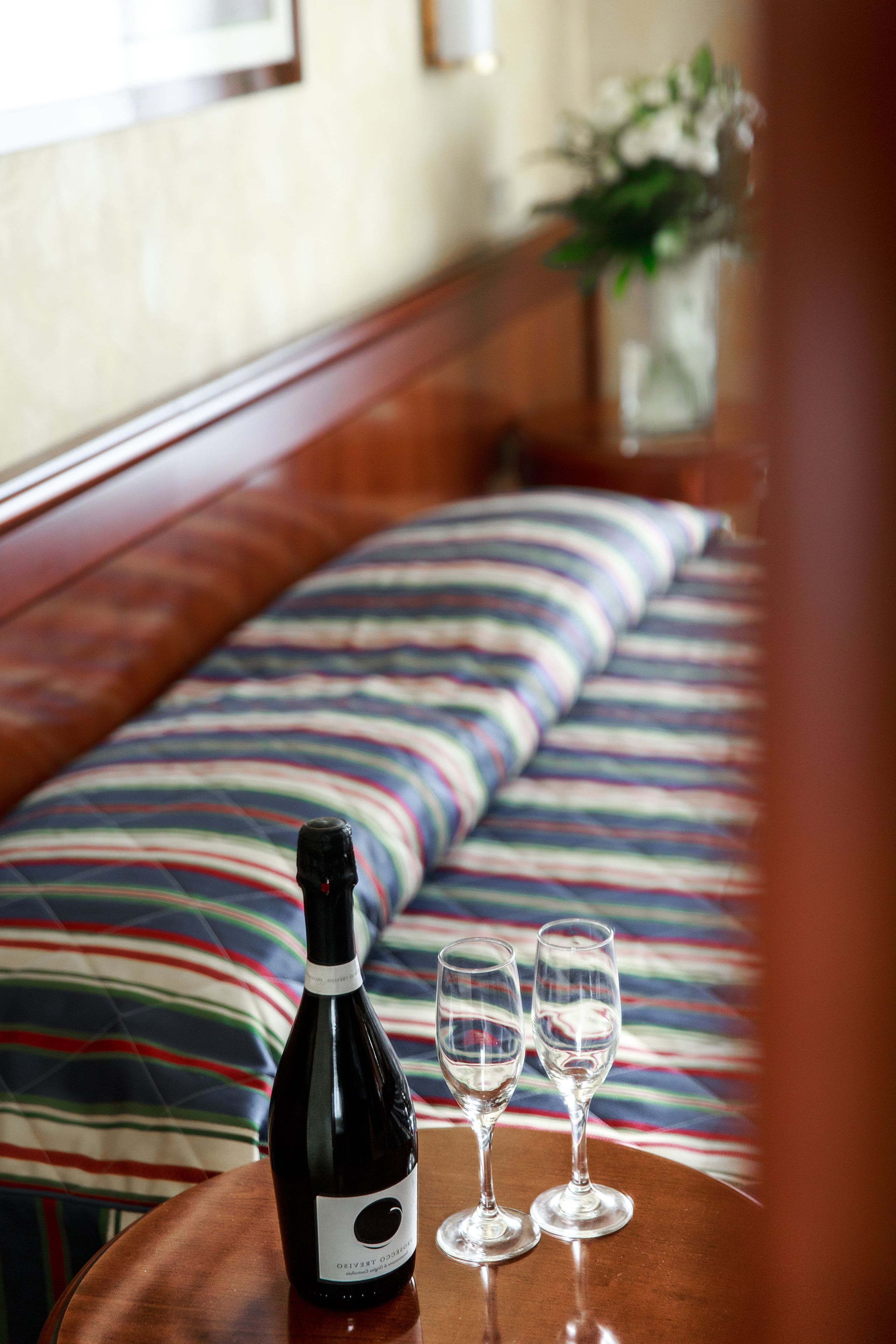 A bed with a colorful striped bedspread, a bottle of wine, and two empty wine glasses on a wooden table.