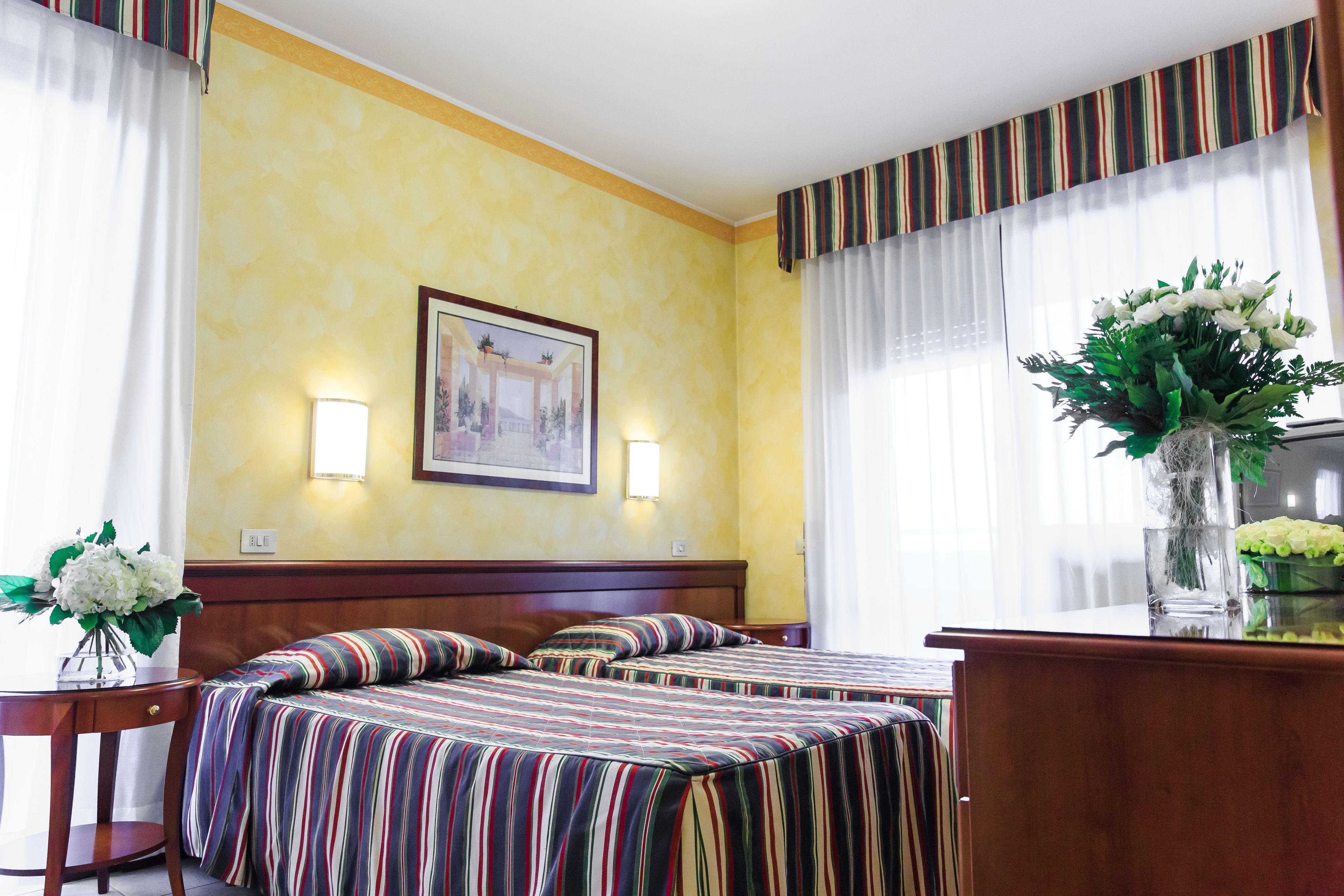 A hotel room with two beds, a wooden headboard, a framed picture on the wall, and a vase with flowers on a table.