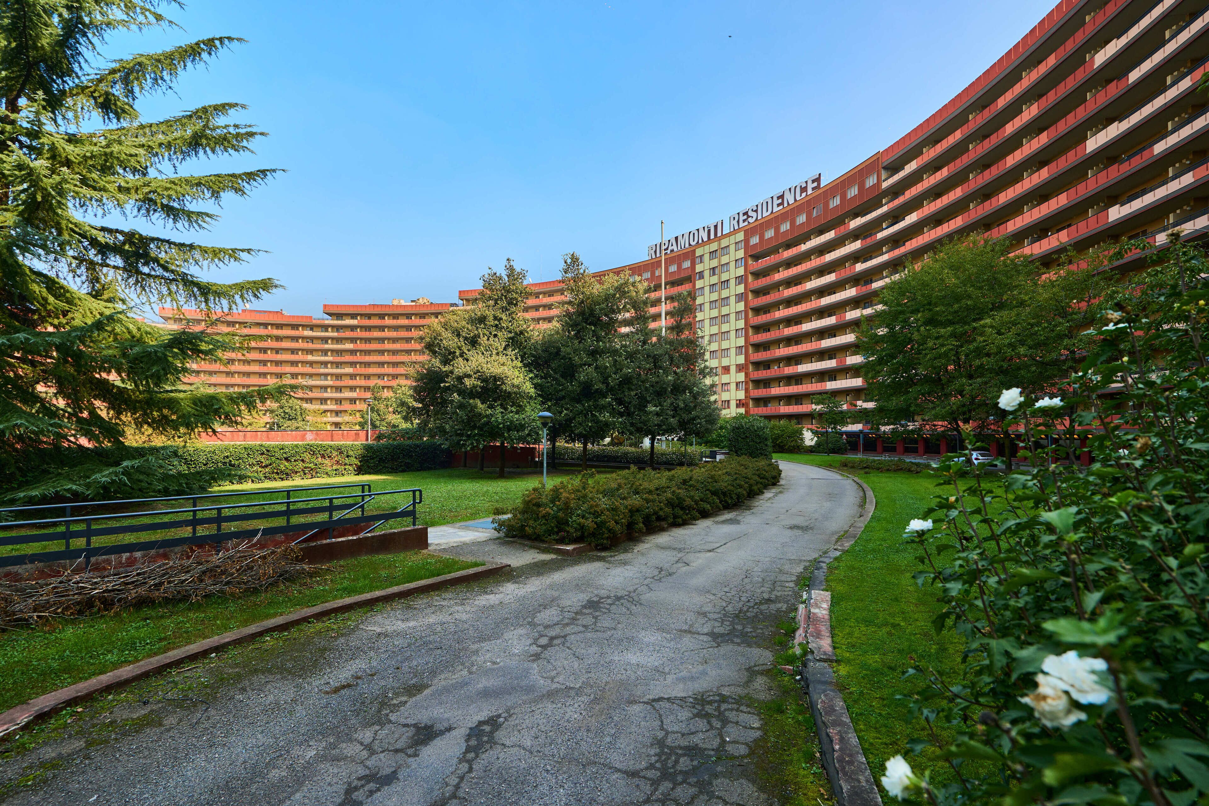 A multi-story building with a curved facade, surrounded by greenery and a paved pathway.