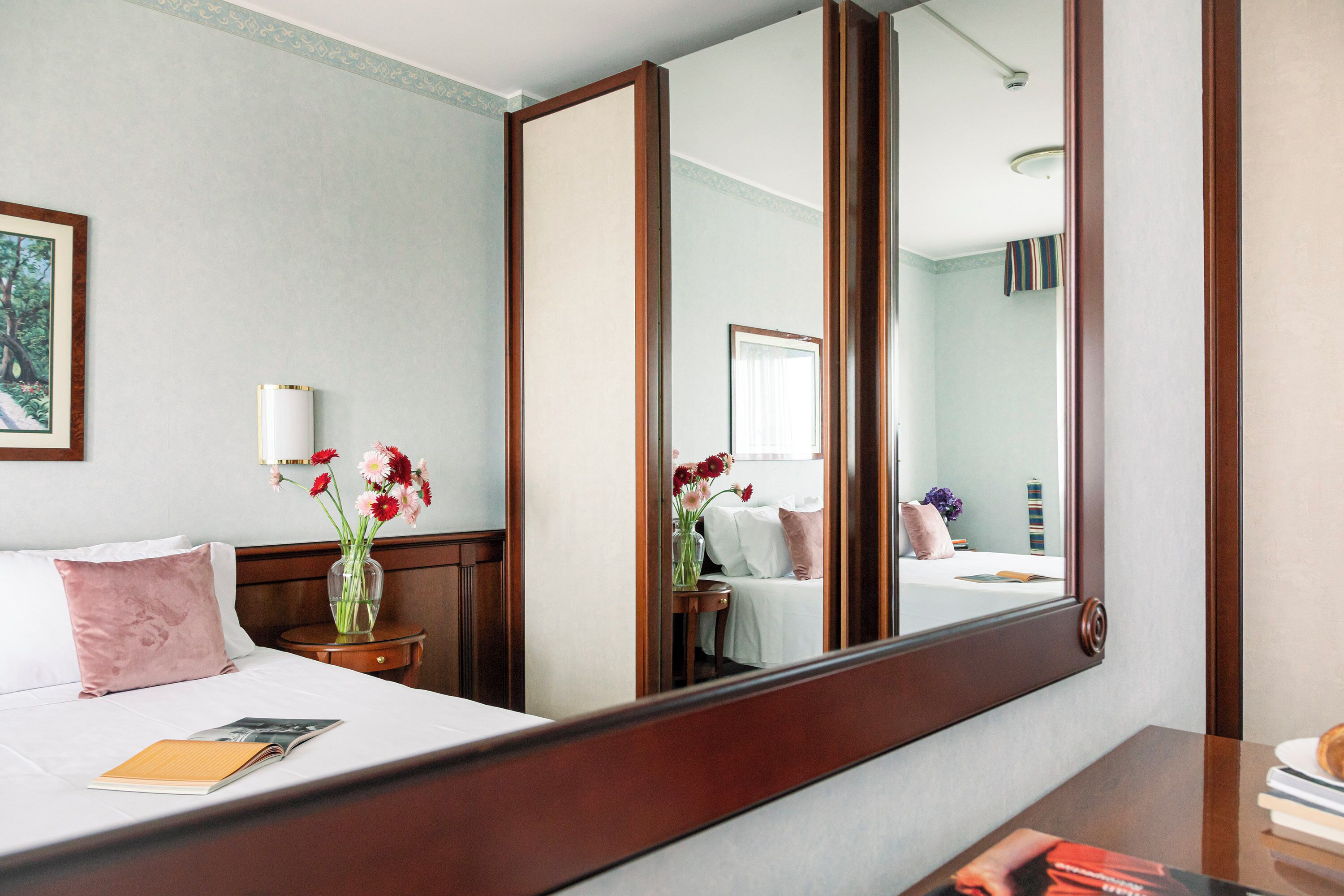 A hotel room with a large mirror, a bed with white linens, a bedside table with a vase of flowers, and a view of the bathroom through the mirror.