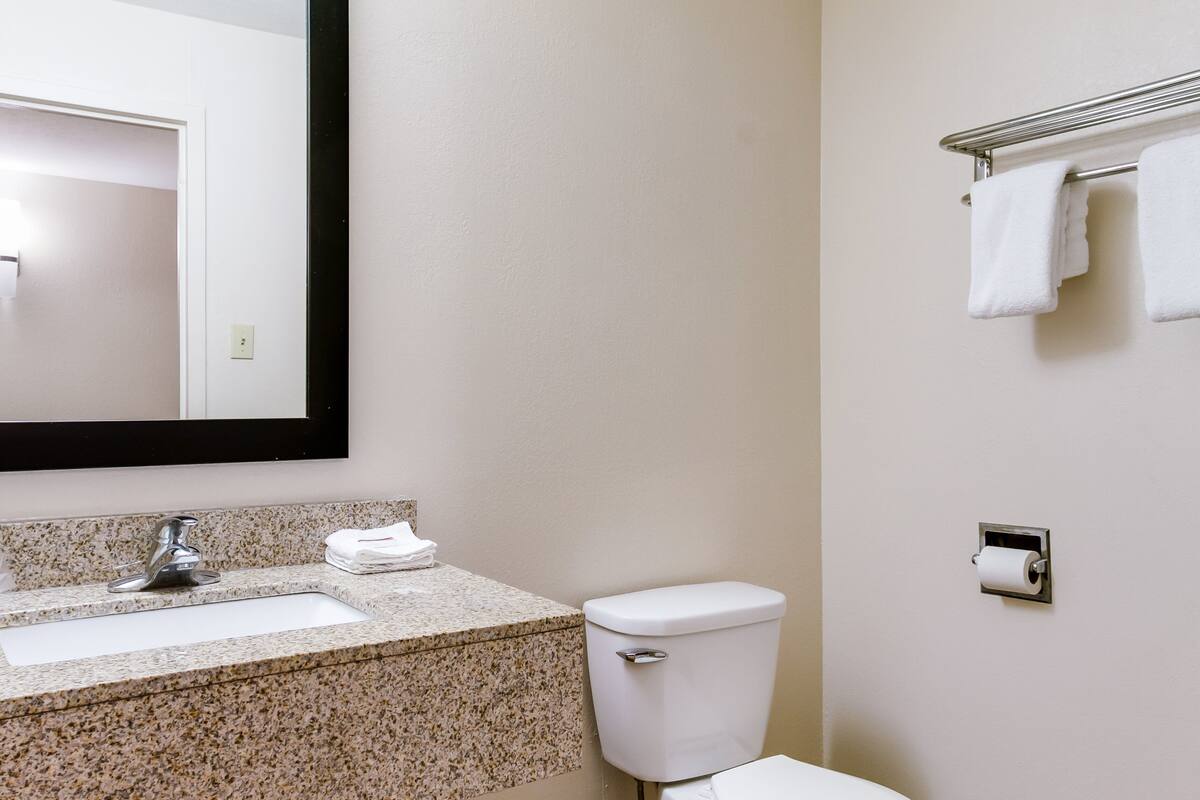 A bathroom with a granite countertop sink, a toilet, a towel rack with towels, and a mirror.