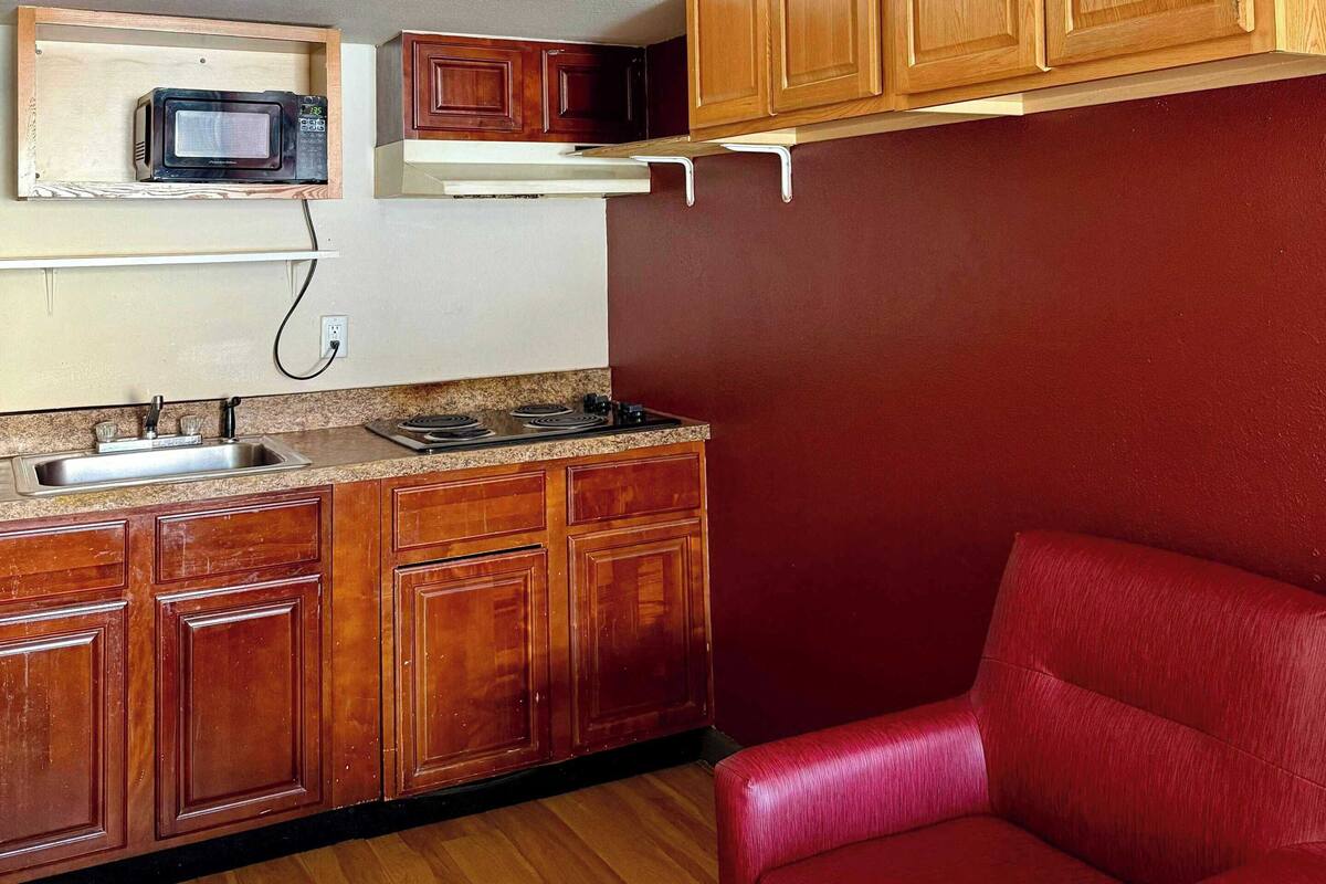 A compact kitchen with wooden cabinets, a microwave, a stove, and a red chair.