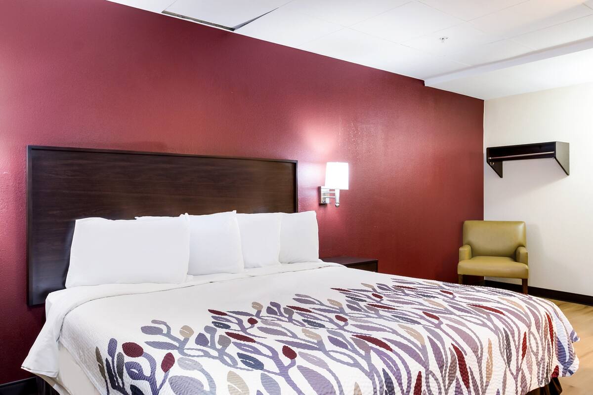 A hotel room with a large bed, a headboard, a wall-mounted lamp, a chair, and a wooden floor.