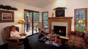 A living room with a fireplace, a sofa, and a chair. There is a person standing by the window and another person sitting on the floor. - The Evergreens On Fall River (Estes Park)