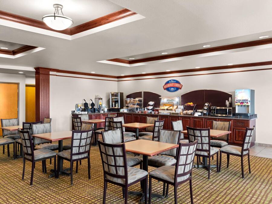 Un espace pour le petit-déjeuner à l’hôtel, avec des tables et des chaises, un comptoir avec une machine à café et un panneau indiquant l’emplacement du buffet du petit-déjeuner.