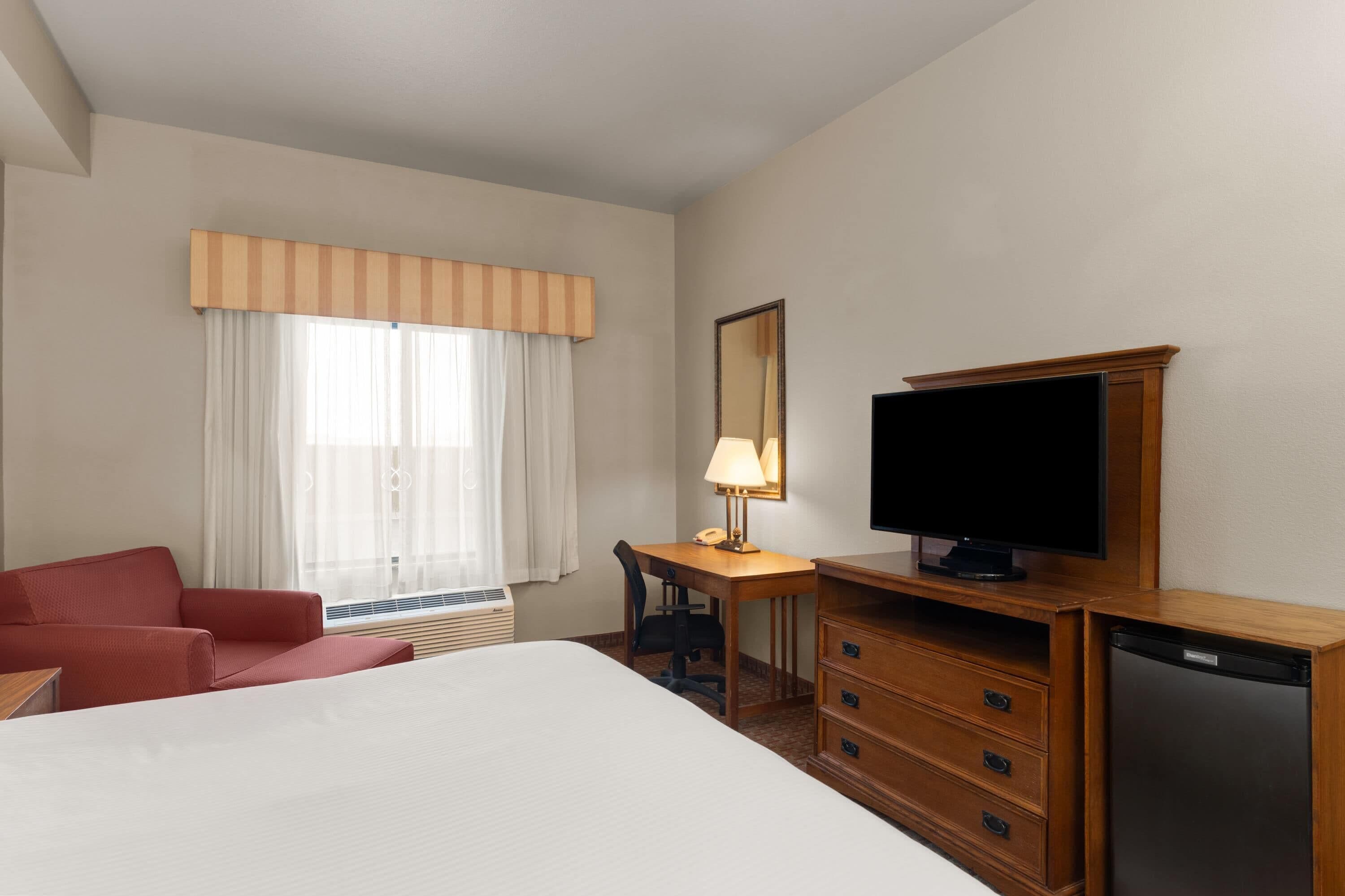A hotel room with a bed, a desk with a lamp, a television, a red chair, and a wooden dresser.