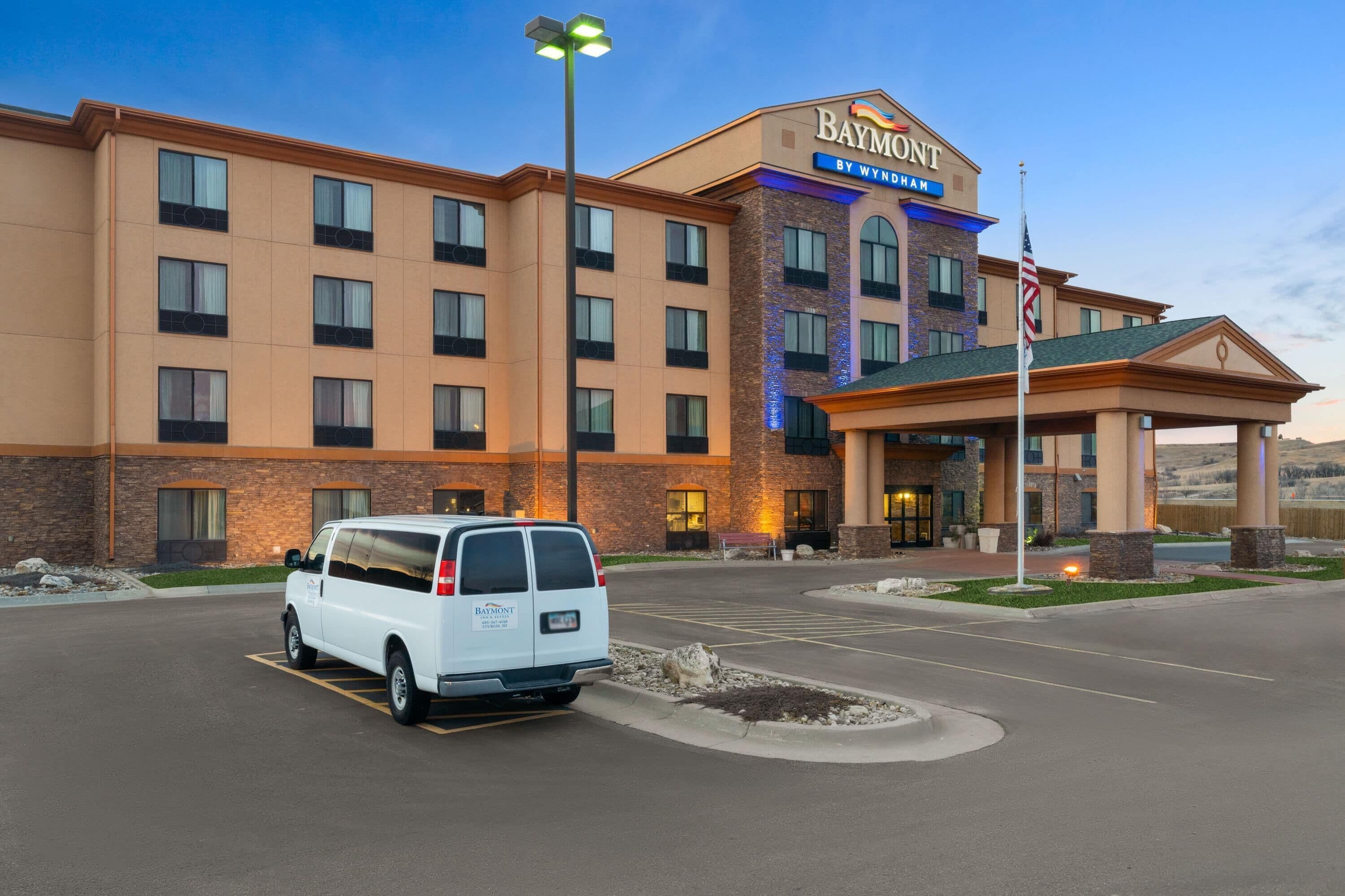 Un hotel Baymont by Wyndham con una camioneta blanca estacionada frente a él, una entrada bien iluminada y una bandera en el asta.