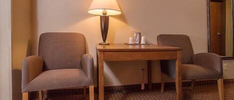 A hotel room with two chairs, a small wooden table, a lamp, and a mirror on the wall.