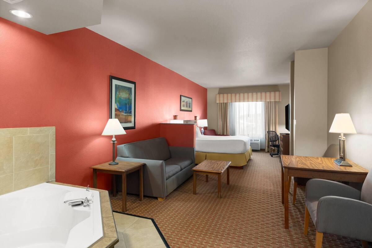 Een hotelkamer met een rode wand, een groot bed, een slaapbank, een bureau, een stoel, een klein tafeltje, een lamp en een bad.
