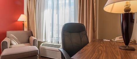 A hotel room with a wooden desk, a black office chair, a striped armchair, a standing lamp, and a window with curtains.