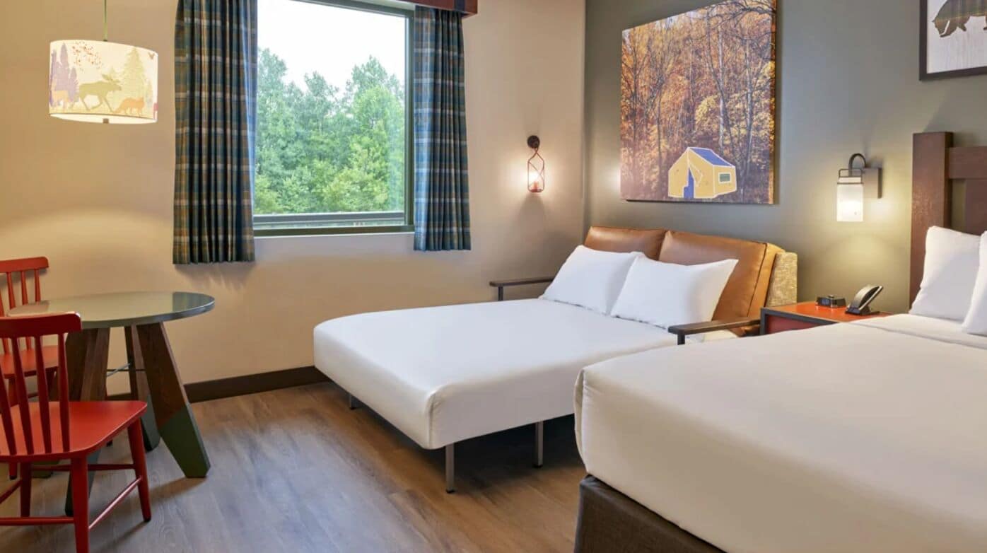 A hotel room with two beds, a small table, a chair, a window with curtains, a painting on the wall, and a lamp.