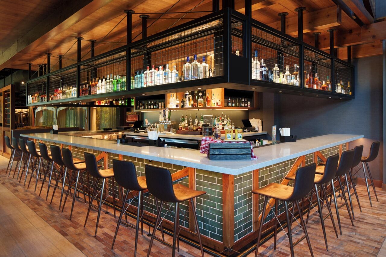 A bar area with a long counter, multiple stools, and shelves stocked with bottles.