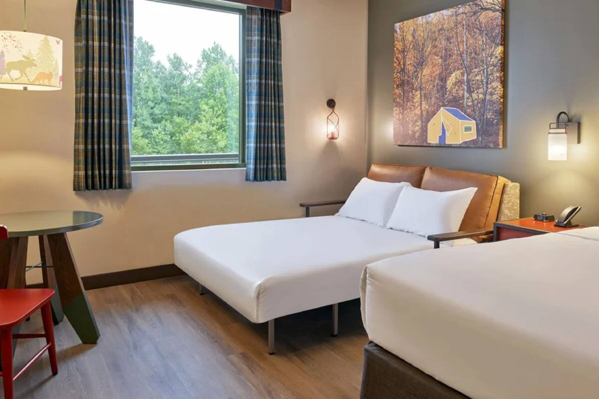 Habitación de hotel con dos camas, una mesita, una silla y un ventanal con cortinas.