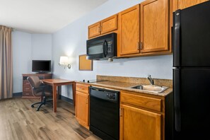 A compact kitchen with wooden cabinets, a microwave, a refrigerator, a sink, and a dishwasher. - Suburban Studios Fort Smith (Fort Smith)