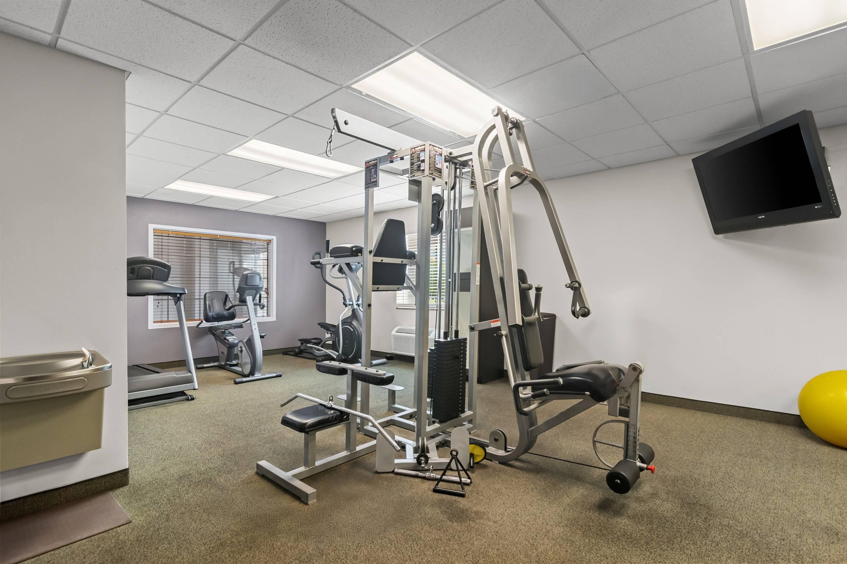 Ein Fitnessraum mit unterschiedlichen Trainingsgeräten, einem Fernseher an der Wand und einem Fenster mit Jalousien.