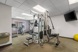A gym with various exercise equipment, a television on the wall, and a window with blinds. - Suburban Studios Fort Smith (Fort Smith)