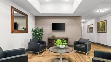 A modern waiting area with leather chairs, a round table with a plant, a flat-screen TV, and a large mirror on the wall.