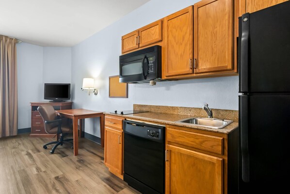 Una cocina compacta con armarios de madera, microondas, refrigerador, lavamanos y un escritorio pequeño con una silla.