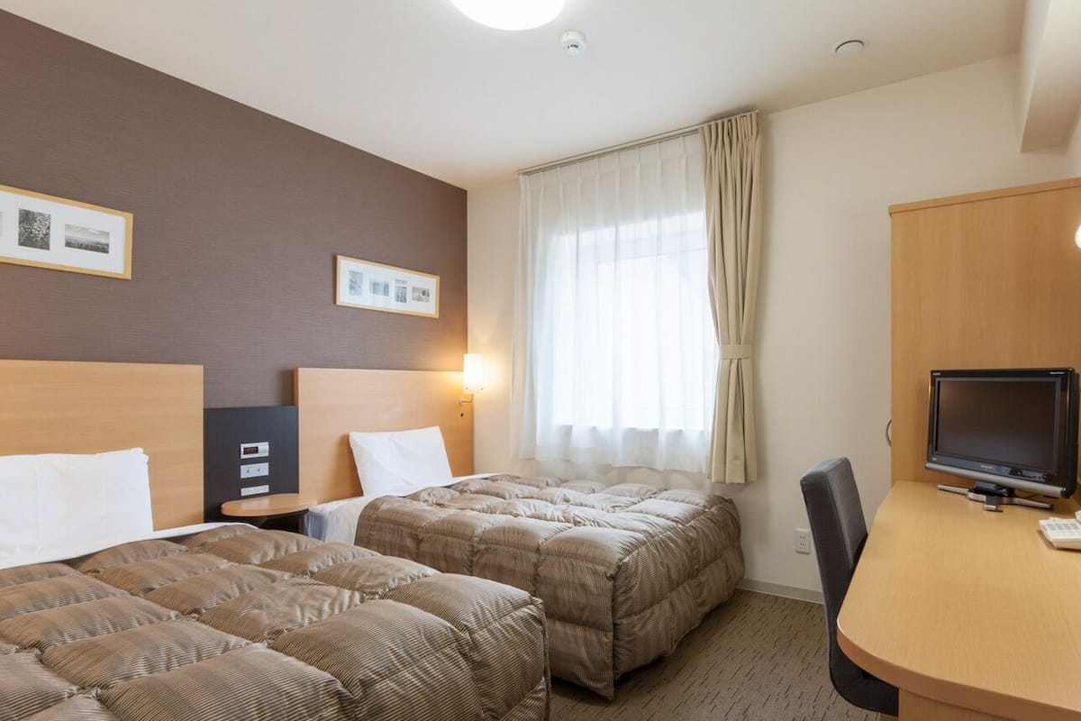 A hotel room with two beds, a desk with a TV, and a window with curtains.