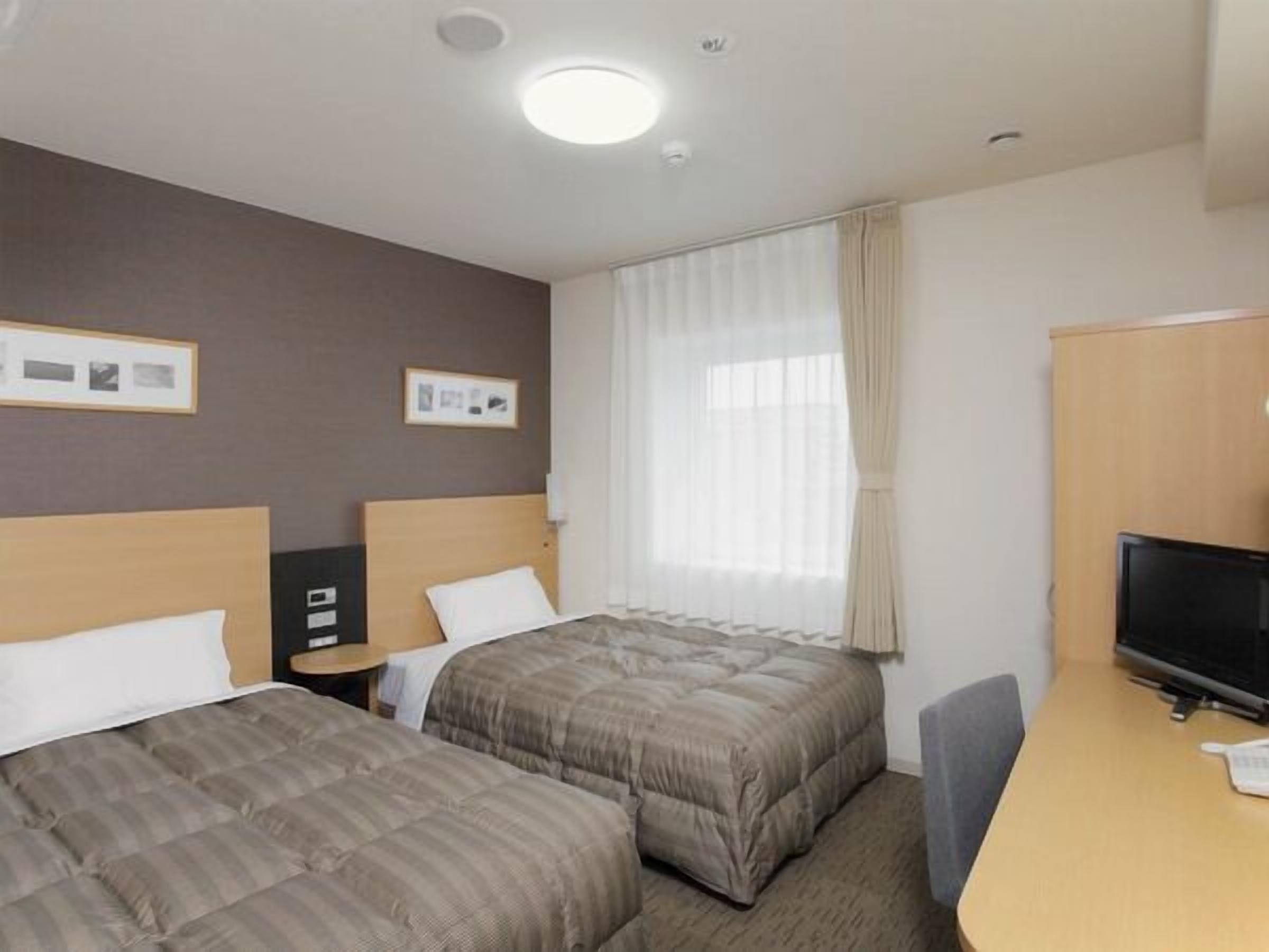 Habitació d'hotel amb dos llits, una taula de treball, un televisor i una finestra amb cortines.