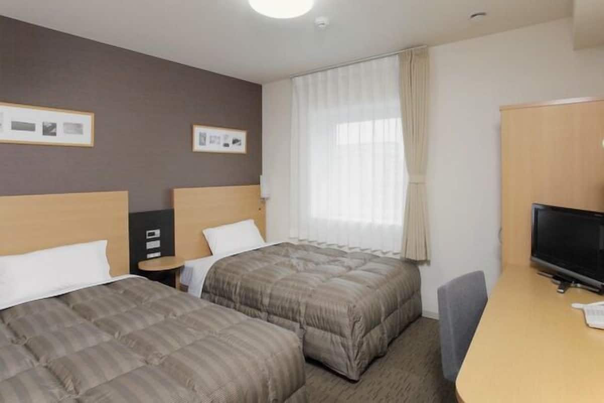 A hotel room with two beds, a desk, a TV, and a window with curtains.