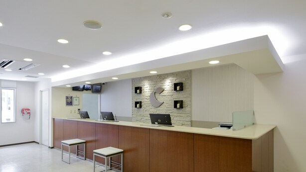 A reception area with a wooden counter, two bar stools, a flat-screen TV, and a stone-patterned wall. - Comfort Hotel Kitami (Kitami)