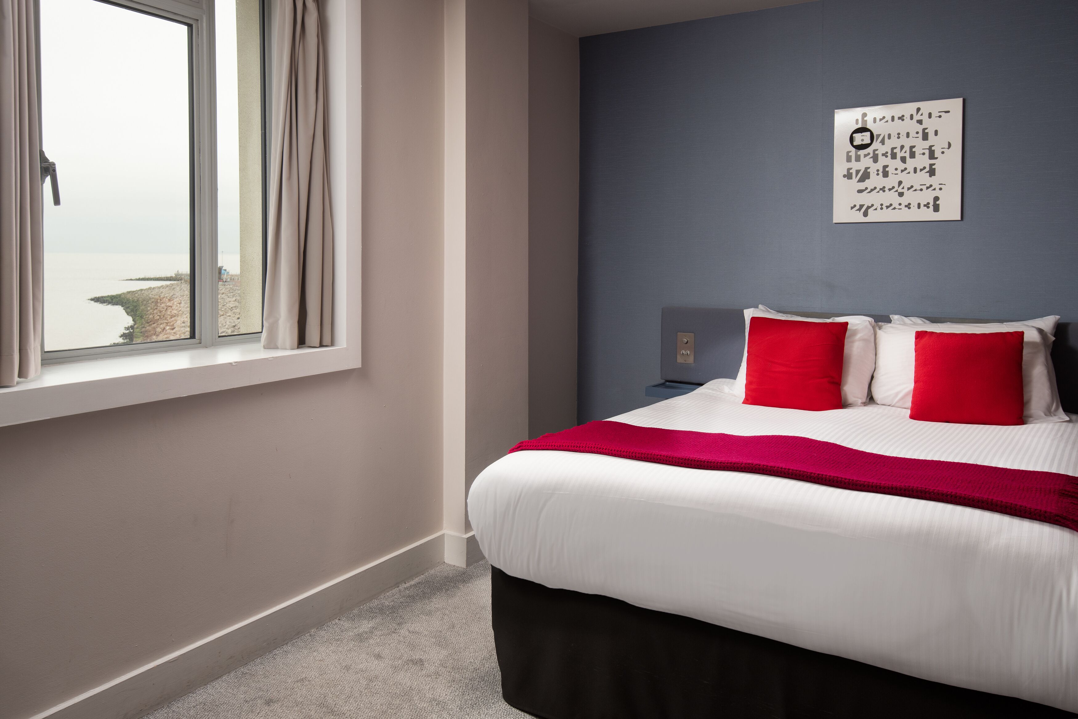Een slaapkamer met een bed, een raam met gordijnen, een aan de muur hangend kunstwerk en een nachtkastje met een lamp.