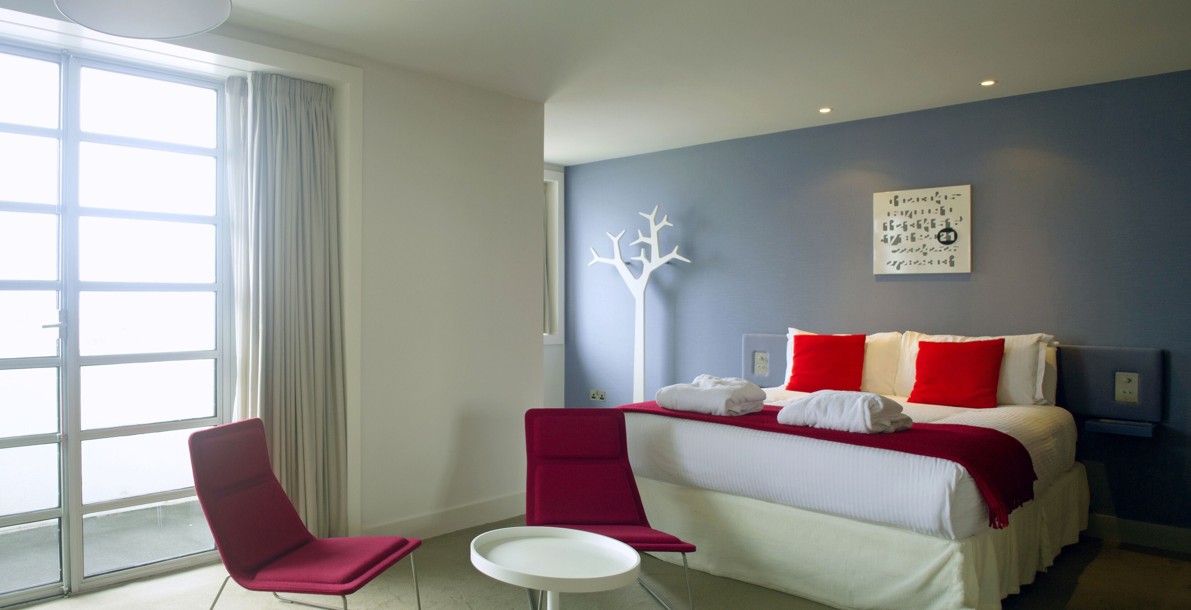 A modern hotel room with a large window, a bed with red and white bedding, a white round table, and a red chair.