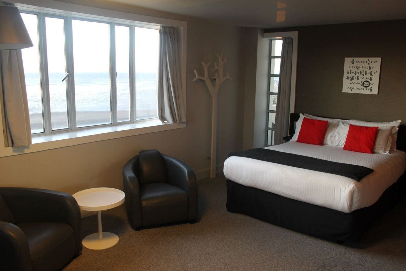 A hotel room with a large bed, a chair, a small table, and a view of the sea through the window.