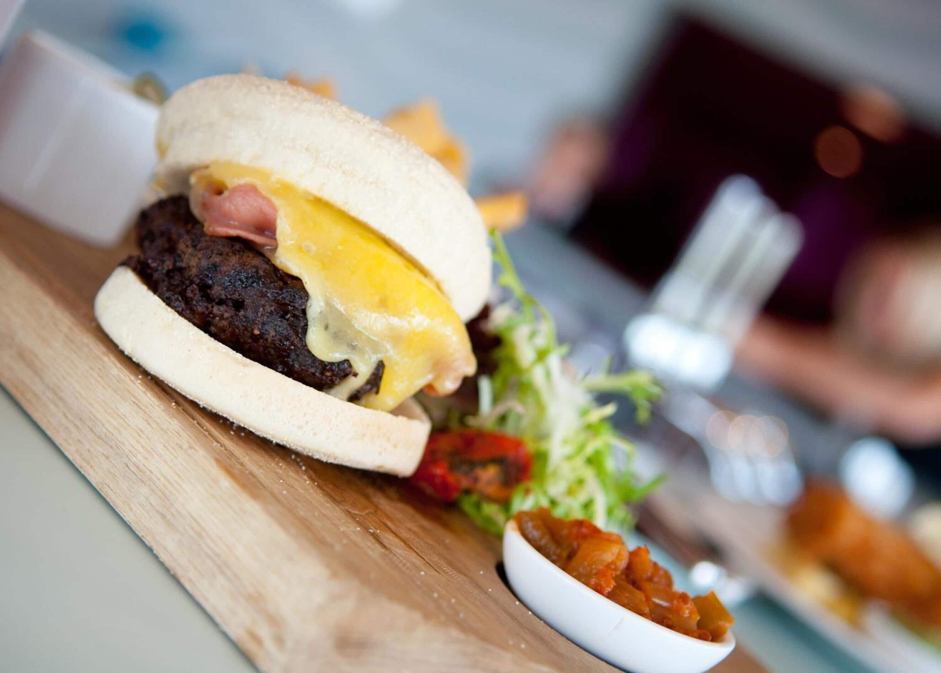 A burger with a beef patty, cheese, and a fried egg on a bun, accompanied by a side salad and a small bowl of pickles.