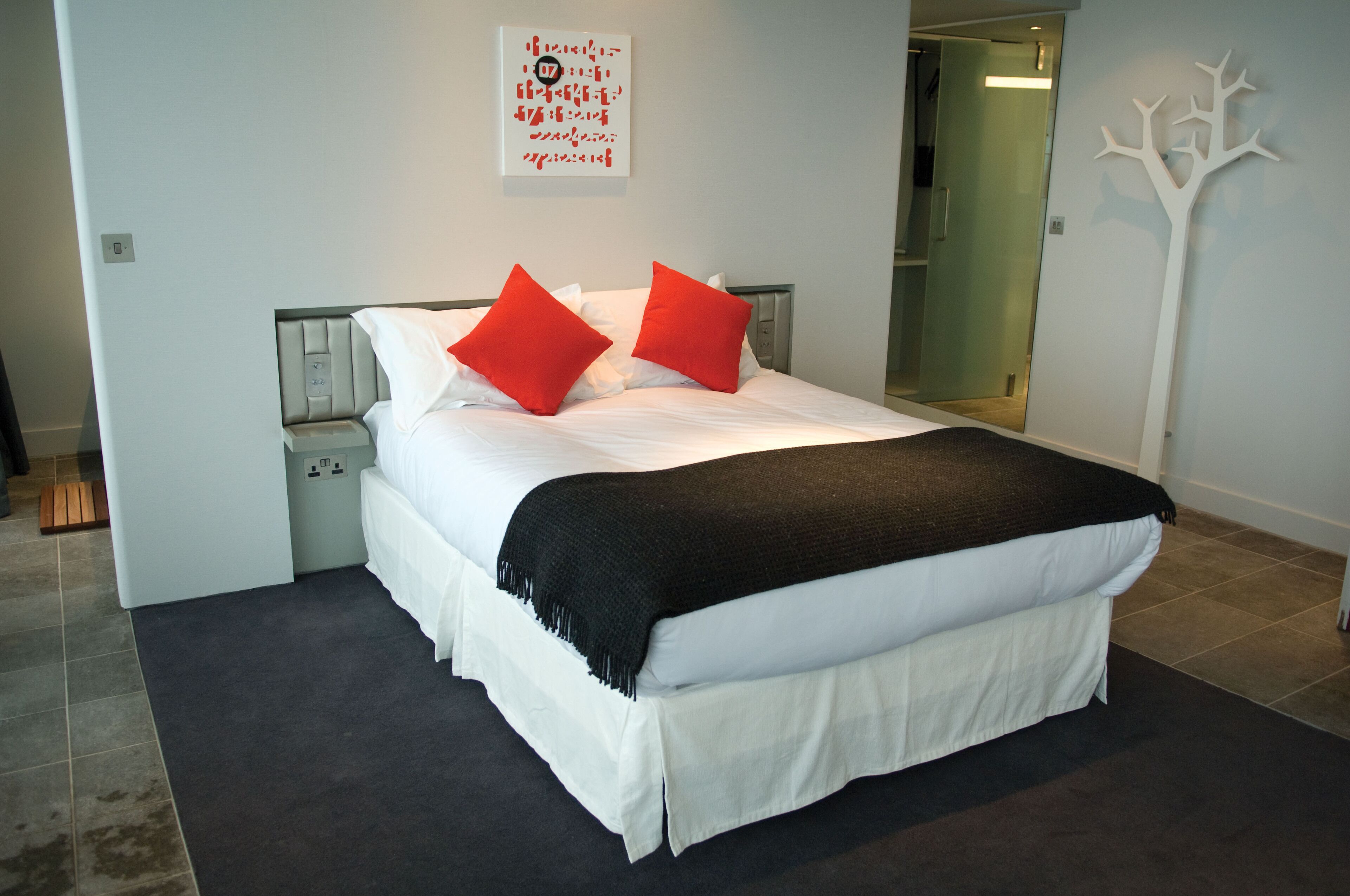 A modern bedroom with a bed, two red pillows, a black throw blanket, and a white wall with a tree decoration.
