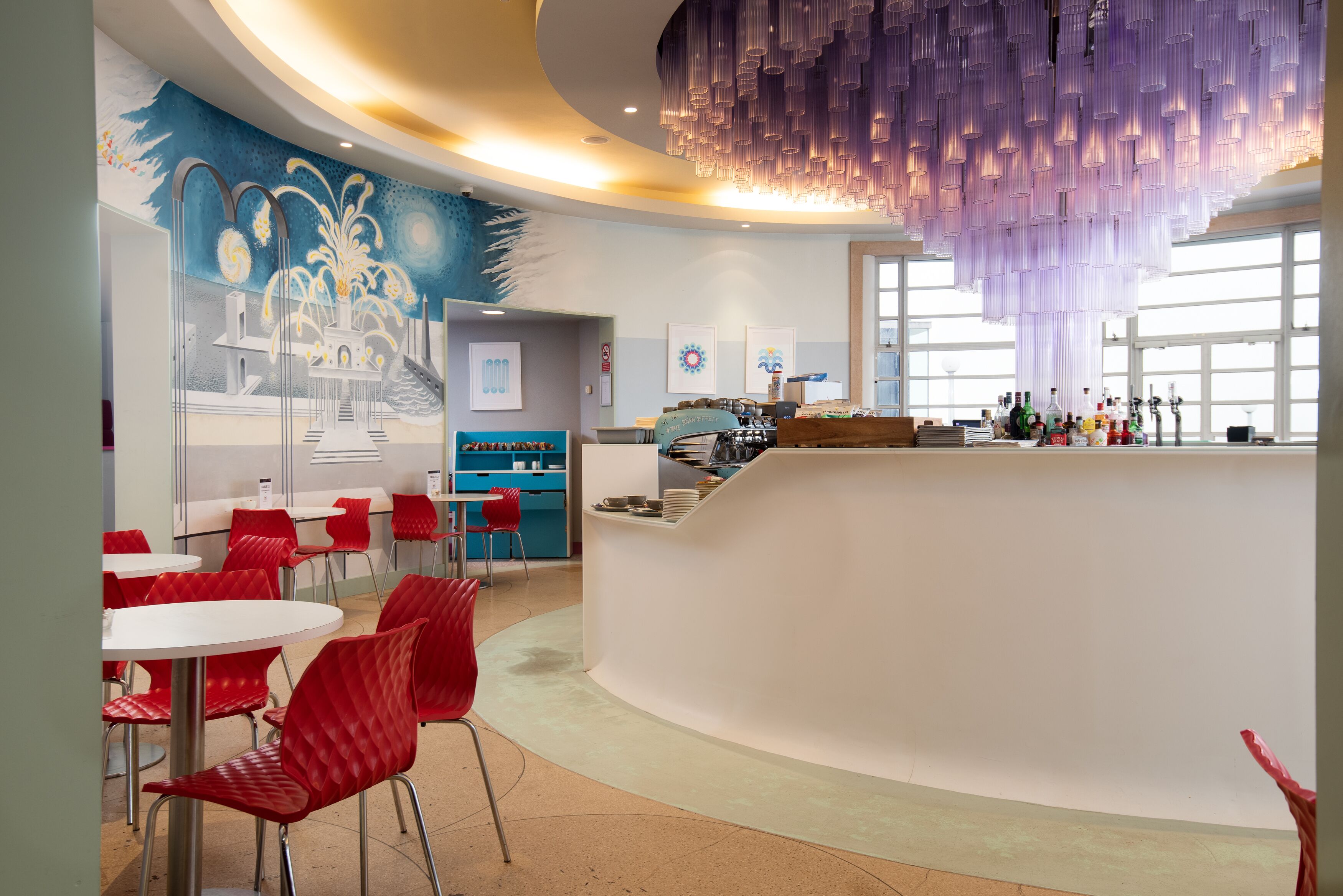 A modern bar area with red chairs, a white counter, and a mural on the wall.