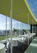 Un restaurante moderno con amplios ventanales, mesas y sillas blancas, y plantas en macetas.