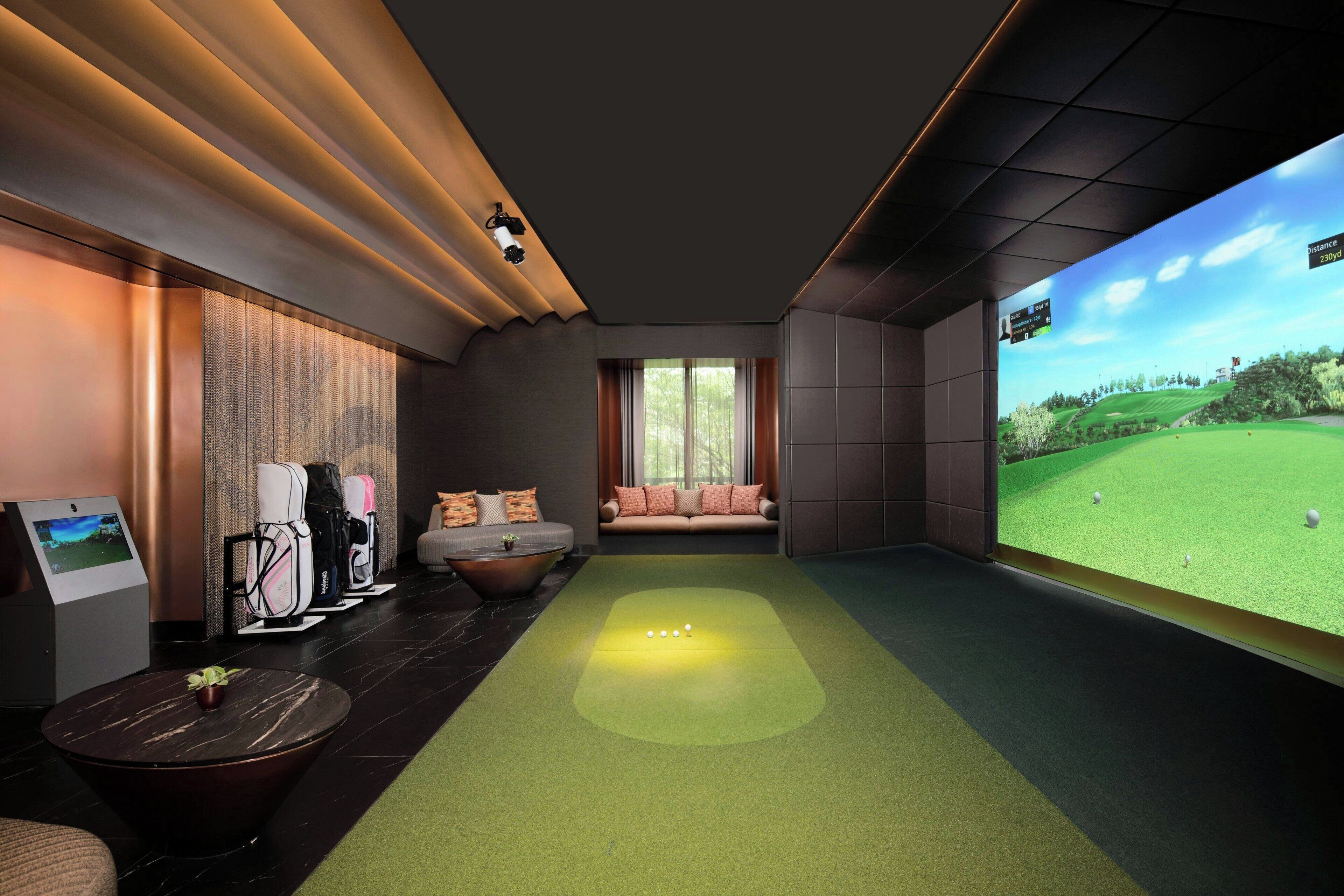 A modern indoor golf simulation room with a large screen displaying a golf course, a green turf area, and a seating area with a sofa and chairs.