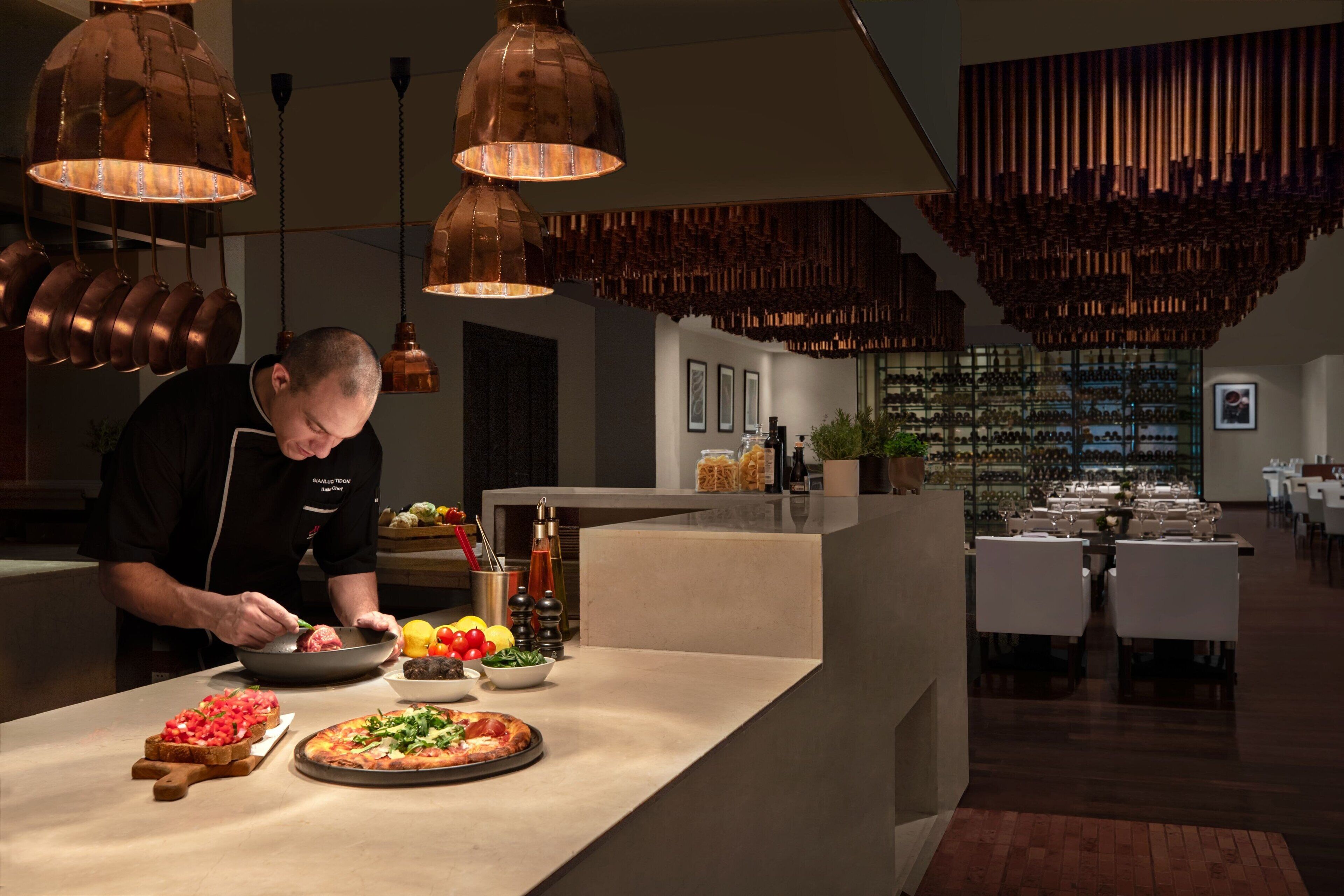 Un chef préparant des plats dans une cuisine moderne, dotée d’éléments en cuivre, avec une cave à vin en arrière-plan.